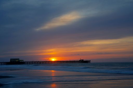 A serene sunset over a pier with vibrant colors reflecting on calm ocean waves, creating a tranquil coastal scene.