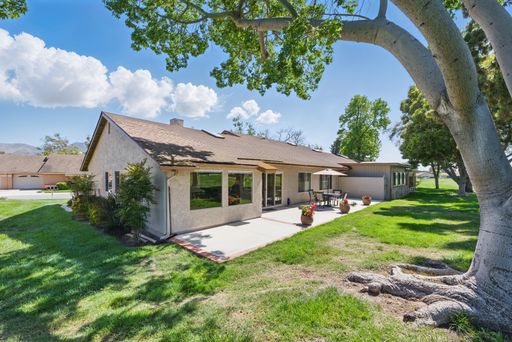 A suburban house with a patio, lush green lawn, and large tree under a bright blue sky with fluffy clouds.