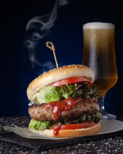 A closeup shot of a hot burger with a cold beer on the table on a dark blue background