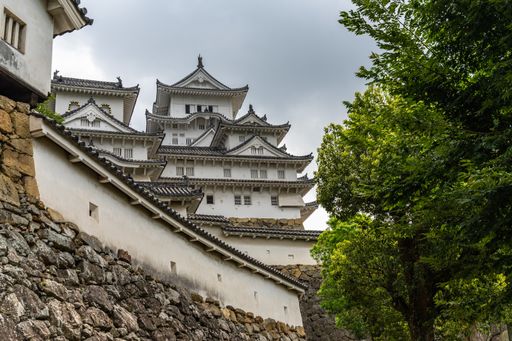 A low angle shot of Himeji Castle, and its stone retaining wall and trees under a cloudy sky in Hyogo, Japan  Himeji Castle Himeji Japan