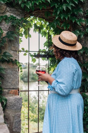 A woman in a hat and dress holding a glass of wine and looking out of the window