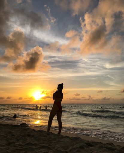 A vertical shot of the silhouette of a female standing on the seacoast under a cloudy sky at sunset