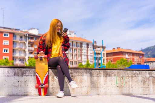 A stylish redhead Hispanic lady using her phone in an urban park holding her skateboard