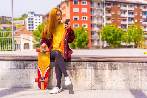 A stylish redhead Hispanic skater lady using her phone enjoying the sunny day in an urban park