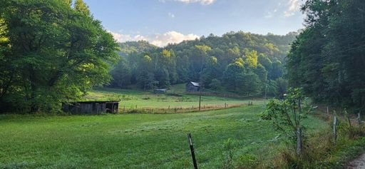 Scenic rural landscape featuring a lush green field, rustic barns, and a backdrop of forested hills under a clear blue sky.