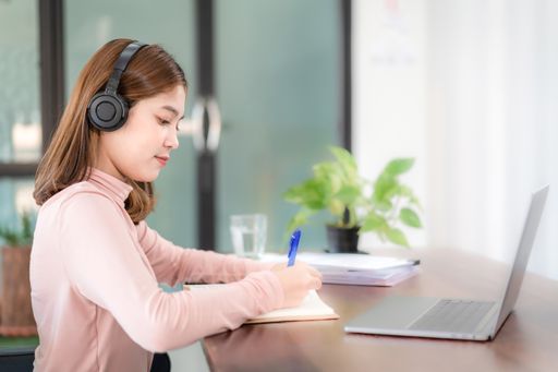 A Southeast Asian college student with headphones writing notes sitting in front of her laptop