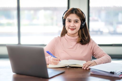 A Southeast Asian college student with headphones writing notes sitting in front of her laptop
