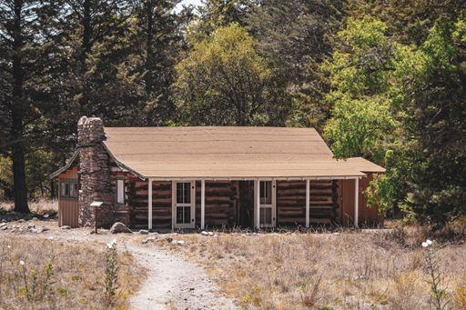 A rustic log cabin surrounded by trees in a serene forest setting, with a dirt path leading to the entrance.