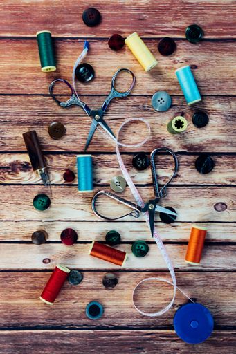 A set of sewing accessories such as spools of colored thread, buttons, measuring tape, vintage scissors, and sewing needles. Top view