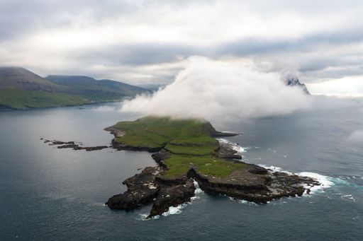 Aerial view of a rugged coastline with lush greenery and clouds, surrounded by the ocean under a cloudy sky.