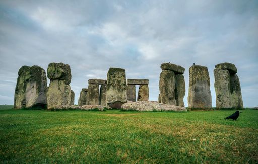 Stonehenge under a cloudy sky with green grass and a crow in the foreground, showcasing ancient stone circle in Wiltshire, England.