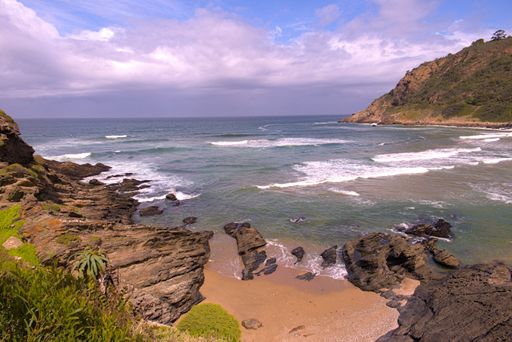 A scenic coastal view with rocky cliffs, sandy beach, and ocean waves under a partly cloudy sky