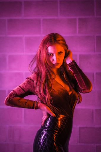 A sexy redhead female in latex tight bodysuit posing in front of a wall illuminated with pink light
