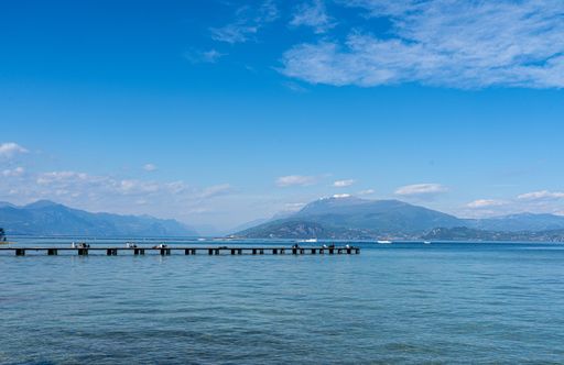 Scenic view of a pier extending into Lake Garda with distant mountains under a clear blue sky, people enjoying the serene setting.