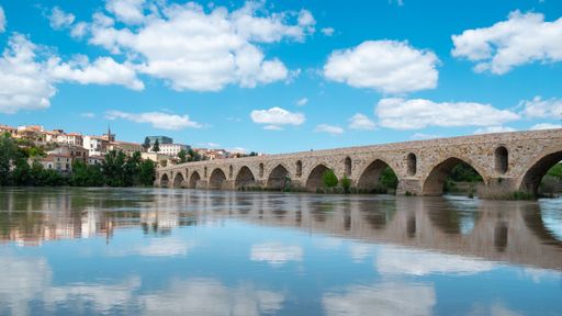 A scenic view of a historic stone bridge over a calm river under a bright blue sky with fluffy clouds in a picturesque town setting.