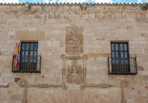 Facade of an ancient stone building with two balconies and Spanish flags, featuring detailed carvings, in a historic European city.
