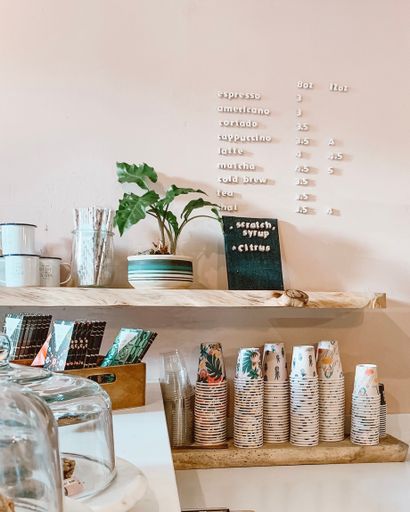 A cozy coffee shop interior with a menu board, potted plant, and stacked colorful cups on wooden shelves.