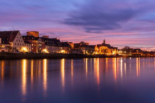 Evening view of Regensburg's historic skyline reflecting in the Danube River, under a vibrant purple and pink sky.