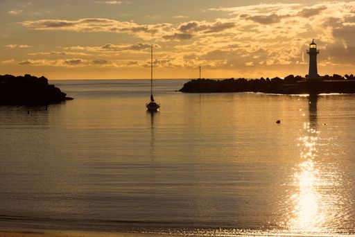 Serene sunset view of a lighthouse and sailboat on calm waters, with golden reflections and a tranquil sky.