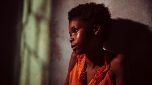 A young African girl lies curled up on her bed, partially wrapped in a worn, patterned blanket. Her face is turned toward the wall, dimly lit by soft,