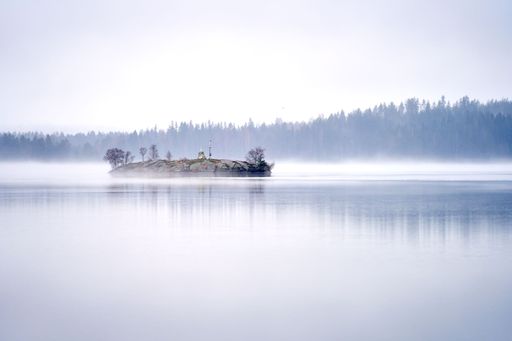 Serene misty lake with a small island and distant forest, creating a tranquil and ethereal landscape.
