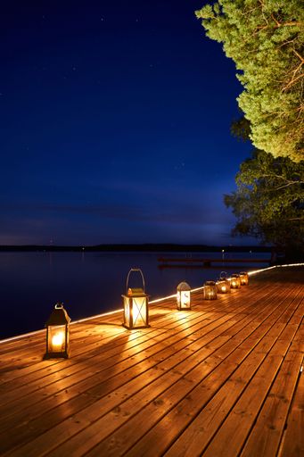 Lanterns illuminate a wooden dock by a calm lake under a starry night sky, creating a peaceful evening atmosphere.