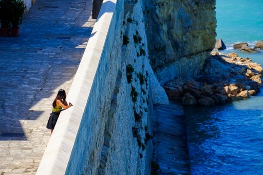 A woman in her 20s leans on a stone wall overlooking a rocky coastline with vibrant blue water on a sunny day.
