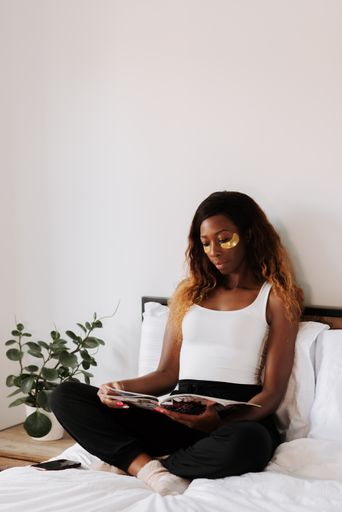 A black beautiful female with anti fatigue under eye masks sitting on the bed reading a journal