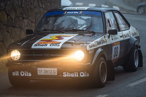 A Ford Escort RS1800 racing in the 73 Rally KH7 Costa Brava, cutting through fog with headlights on, showcasing vintage motorsport heritage