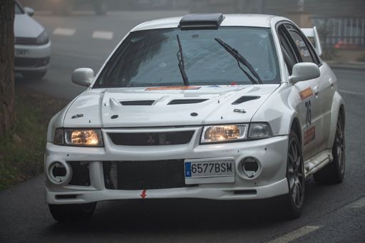 A white Mitsubishi Carisma GT Evo VI rally car with headlights on, driving through foggy conditions on a wet road, showcasing speed, performance