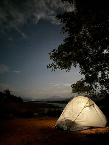 Night camping scene with an illuminated tent under a starry sky, surrounded by nature and a mountain silhouette in the background.