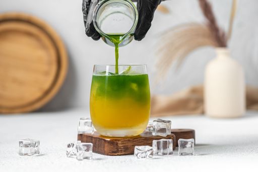 A green drink is poured into a glass with orange drink and ice cubes nearby.