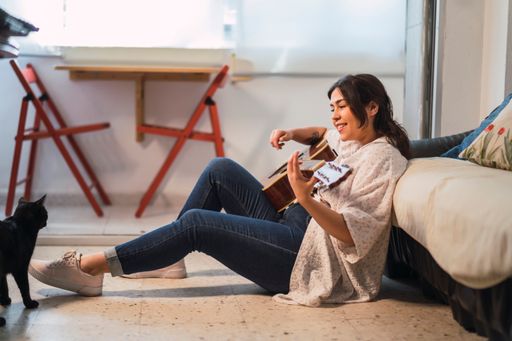A young Caucasian female playing the guitar sitting on the floor and leaning on the couch
