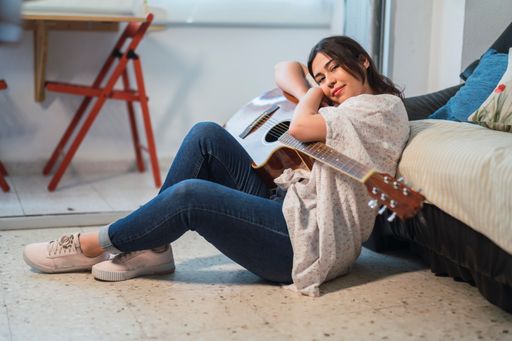 A young Caucasian female playing the guitar sitting on the floor and leaning on the couch