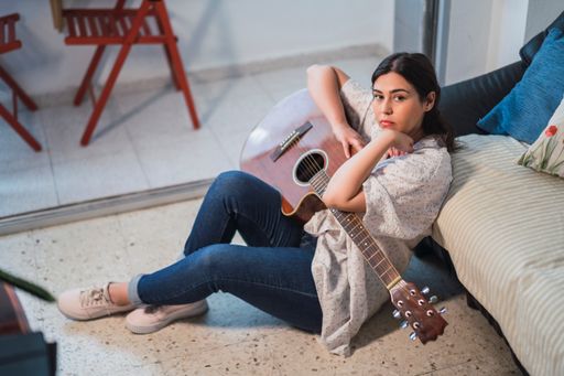 A young Caucasian female playing the guitar sitting on the floor and leaning on the couch