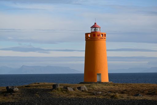 A vibrant orange lighthouse stands on a coastal landscape with distant mountains under a partly cloudy sky in Iceland.