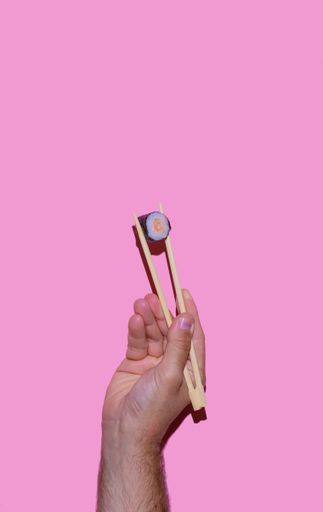 A vertical shot of a hand of a man holding Japanese chopsticks with sushi on a pink background