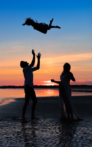 A vertical shot of a father silhouette with his pregnant wife, throwing his child into the air at sunset on the sea
