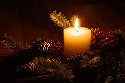 A closeup of a lit candle, surrounded by pine cones in a dark Christmas setting