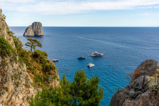 Scenic view of yachts on the blue waters of Capri with lush cliffs and the Faraglioni rock formations under a clear sky.