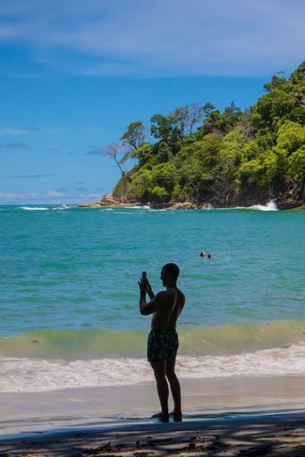 A man in swim shorts takes a photo on a tropical beach with lush greenery and clear blue waters, under a bright sky.