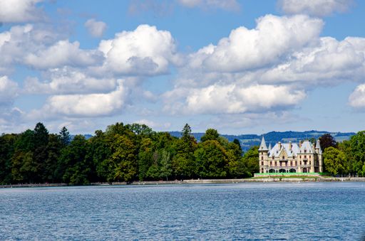 A scenic view of a historic castle by Lake Geneva with lush greenery and a bright blue sky, showcasing European architecture.