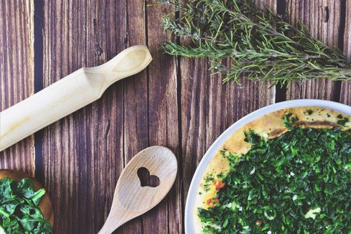 A top view shot of a wooden table with cooked spinach, rosemary on the side, and kitchen utensils