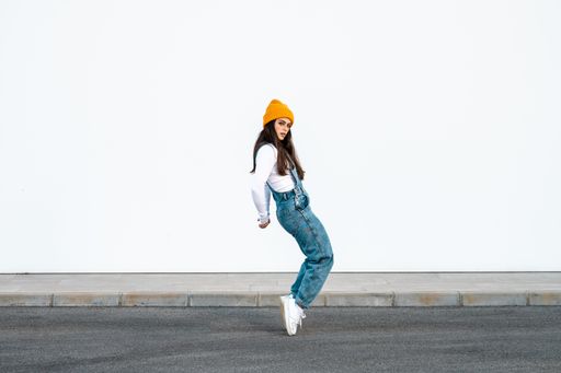 A young Caucasian female wearing denim pants, white shirt and a yellow hat posing against a white wall