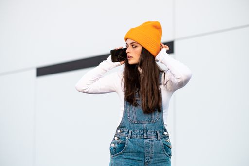 A Caucasian female in denim pants, white shirt and a yellow hat using a phone against a white wall