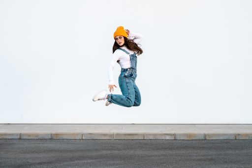 A young Caucasian female wearing denim pants, white shirt and a yellow hat posing against a white wall