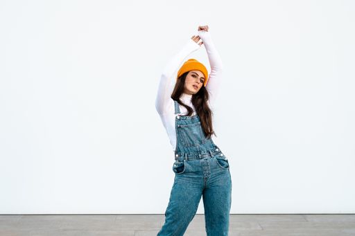 A young Caucasian female wearing denim pants, white shirt and a yellow hat posing against a white wall