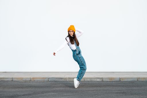A young Caucasian female wearing denim pants, white shirt and a yellow hat posing against a white wall