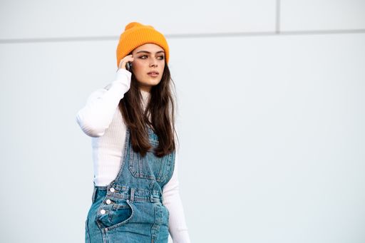 A Caucasian female in denim pants, white shirt and a yellow hat using a phone against a white wall