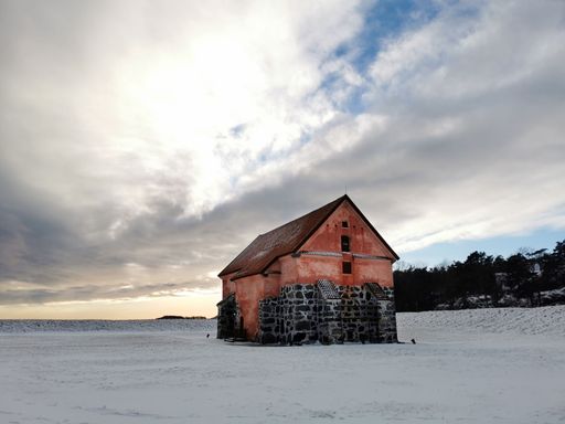 A solitary old barn with stone foundation stands on a snowy field under a dramatic cloudy sky, evoking a serene winter scene.